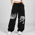 A woman modeling black sweatpants with white skeleton hand and leg graphics, paired with a white crop top and white sneakers, against a light gray studio background.