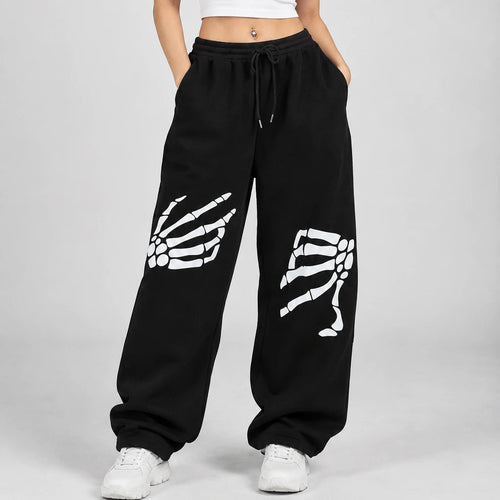 A woman modeling black sweatpants with white skeleton hand and leg graphics, paired with a white crop top and white sneakers, against a light gray studio background.